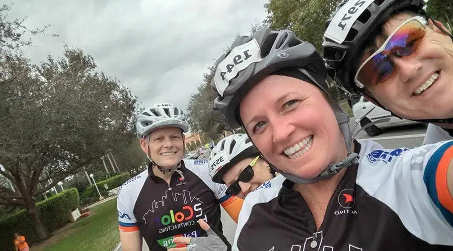Four cyclists wearing helmets and cycling jerseys smile for a group selfie outdoors, with cloudy skies and trees in the background.