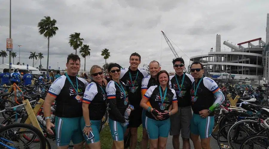 A group of cyclists in jerseys and medals pose together in front of bicycles and a stadium on a cloudy day.