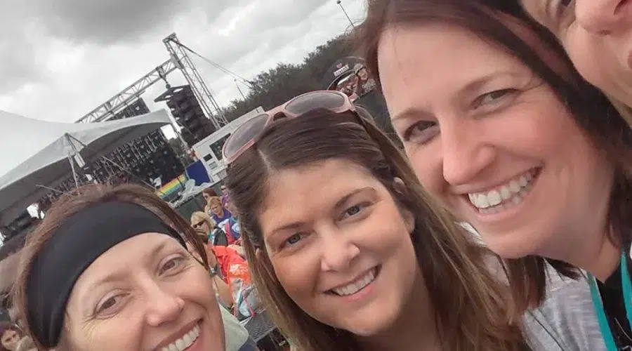 Four women smiling at an outdoor event with a stage and tents in the background.