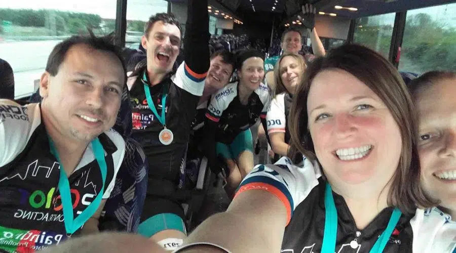 A group of smiling cyclists wearing medals and team jerseys take a selfie while sitting in a bus.