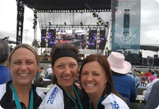 Three women smiling and standing together at an outdoor concert with a stage and banner in the background. The banner displays logos for Pepsi and Hard Rock. Crowd members are seated behind them.