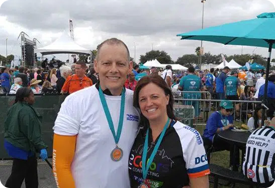 Two people wearing medals pose together at an outdoor event with tents and a crowd in the background. The man is wearing a white shirt and orange sleeves, and the woman is wearing a white and blue jersey.