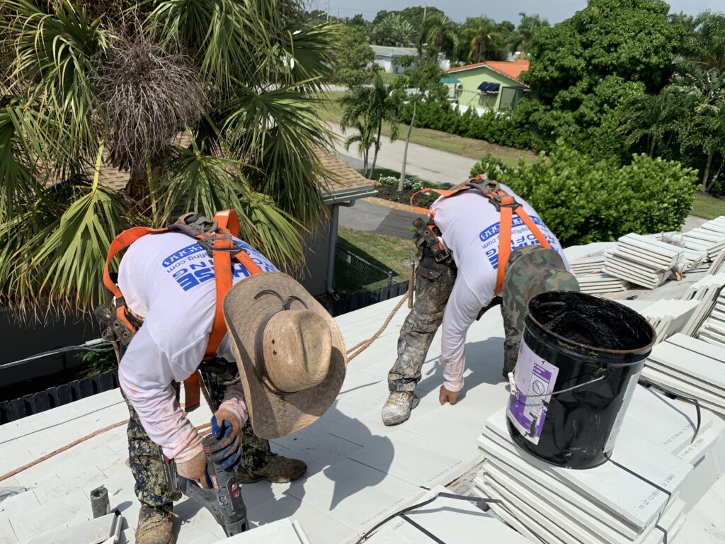 Two construction workers in safety gear are working on a house roof amidst palm trees and residential area. One is adjusting roof tiles while the other handles a black bucket.