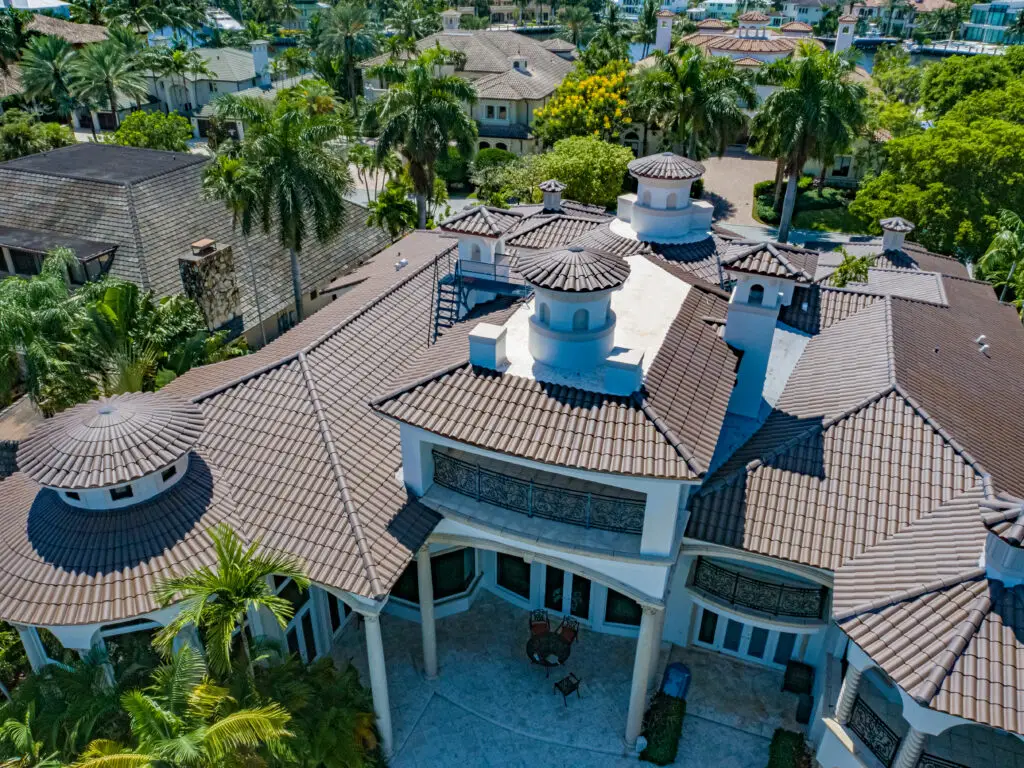 Aerial view of a large, Mediterranean-style roof repair with a brown tile roof, surrounded by palm trees and neighboring homes.