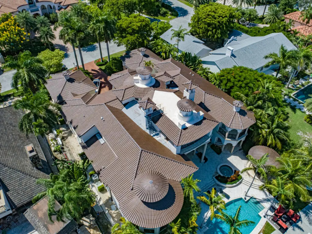 Aerial view of a large, multi-story house with a complex roof design, surrounded by palm trees. Visible backyard features a swimming pool and landscaped garden. Neighboring houses are also seen.