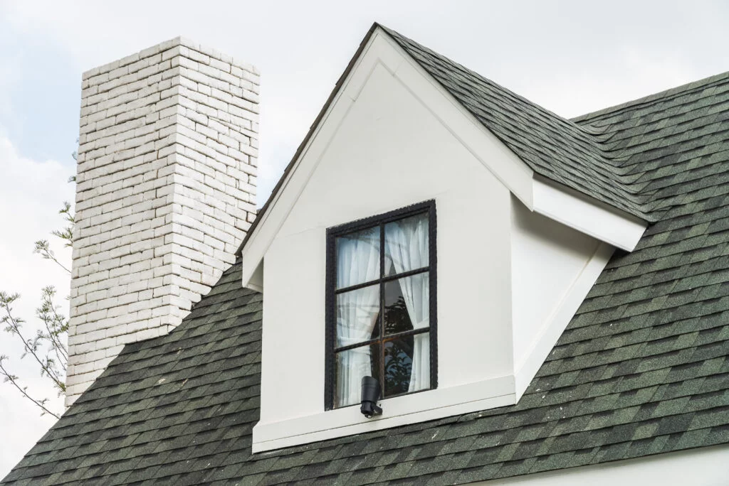 A house roof with class four shingles in green, featuring a white dormer window and a tall brick chimney, ensures impact-resistant roofing.
