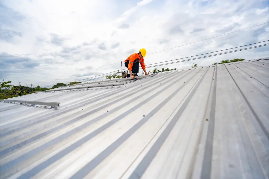 A worker in safety gear installs or repairs a metal roof under a cloudy sky, using tools and positioned near the roof’s peak—a task expertly handled by leading commercial roofing companies near me.